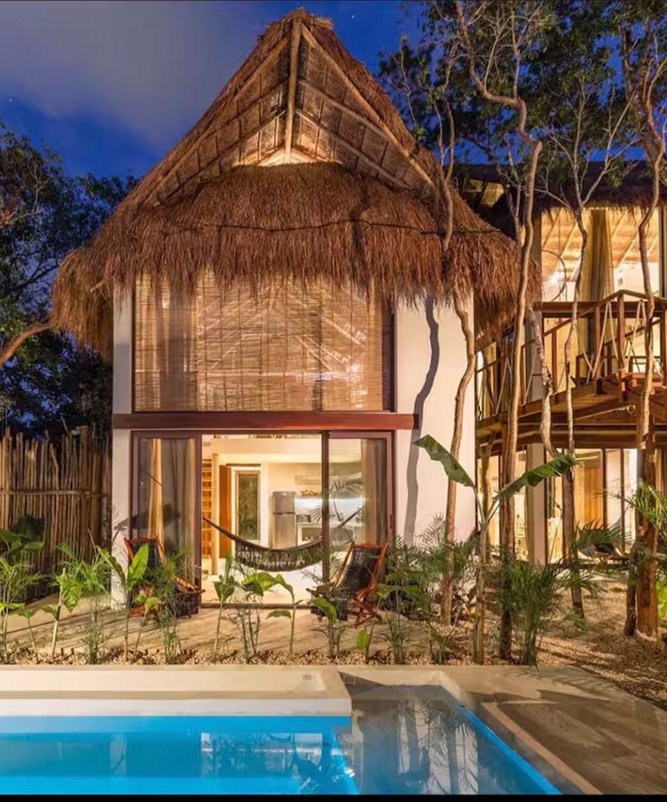 Ikal bungalow with a palm-thatched roof and a lit pool at dusk